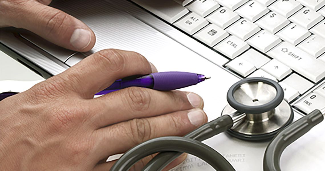 Someone working on the computer, pen in hand, and a stethoscope nearby.